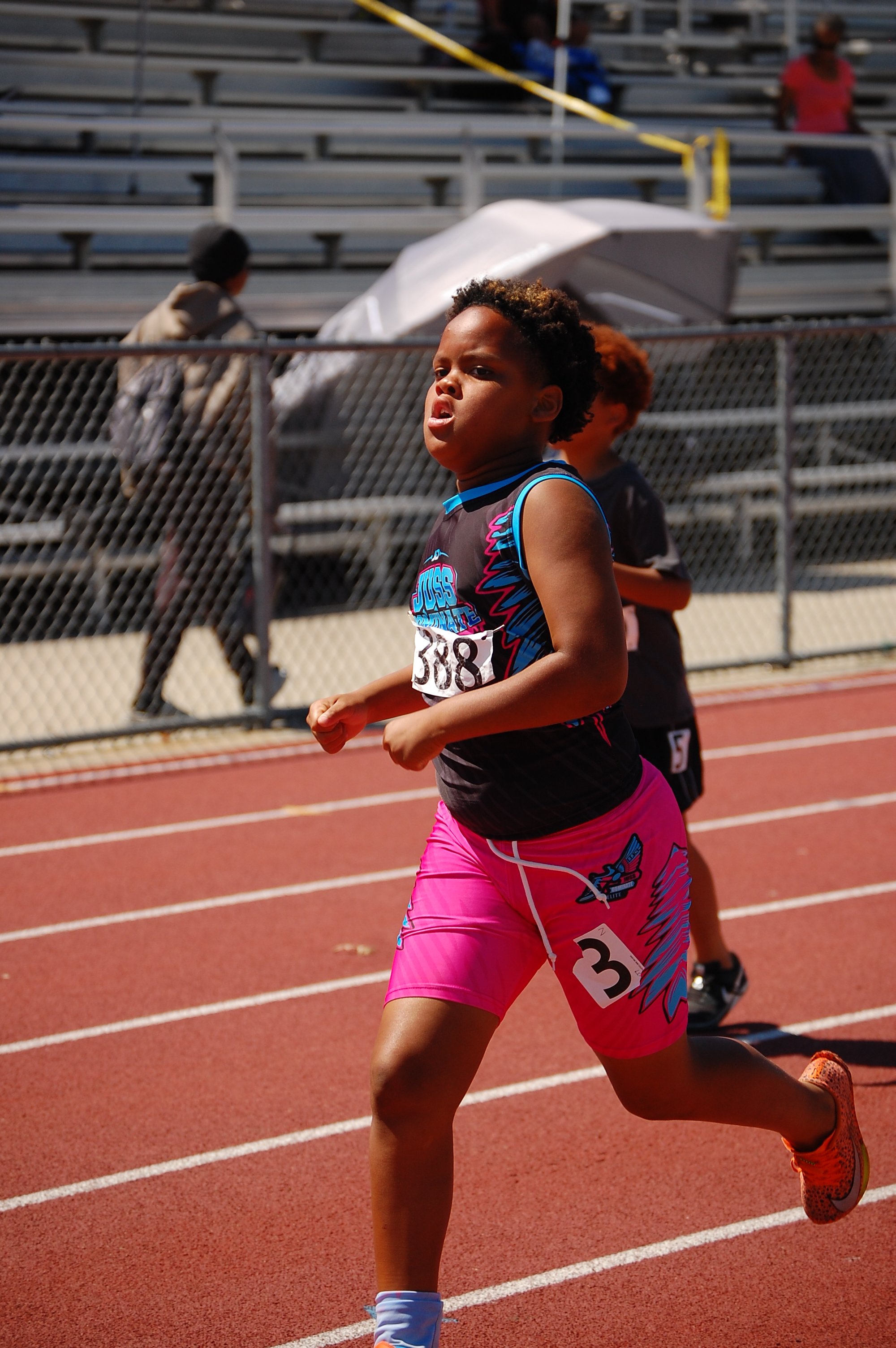 A young female athlete running on a track during a race, wearing a black and pink athletic outfit with the number 38 on her bib.