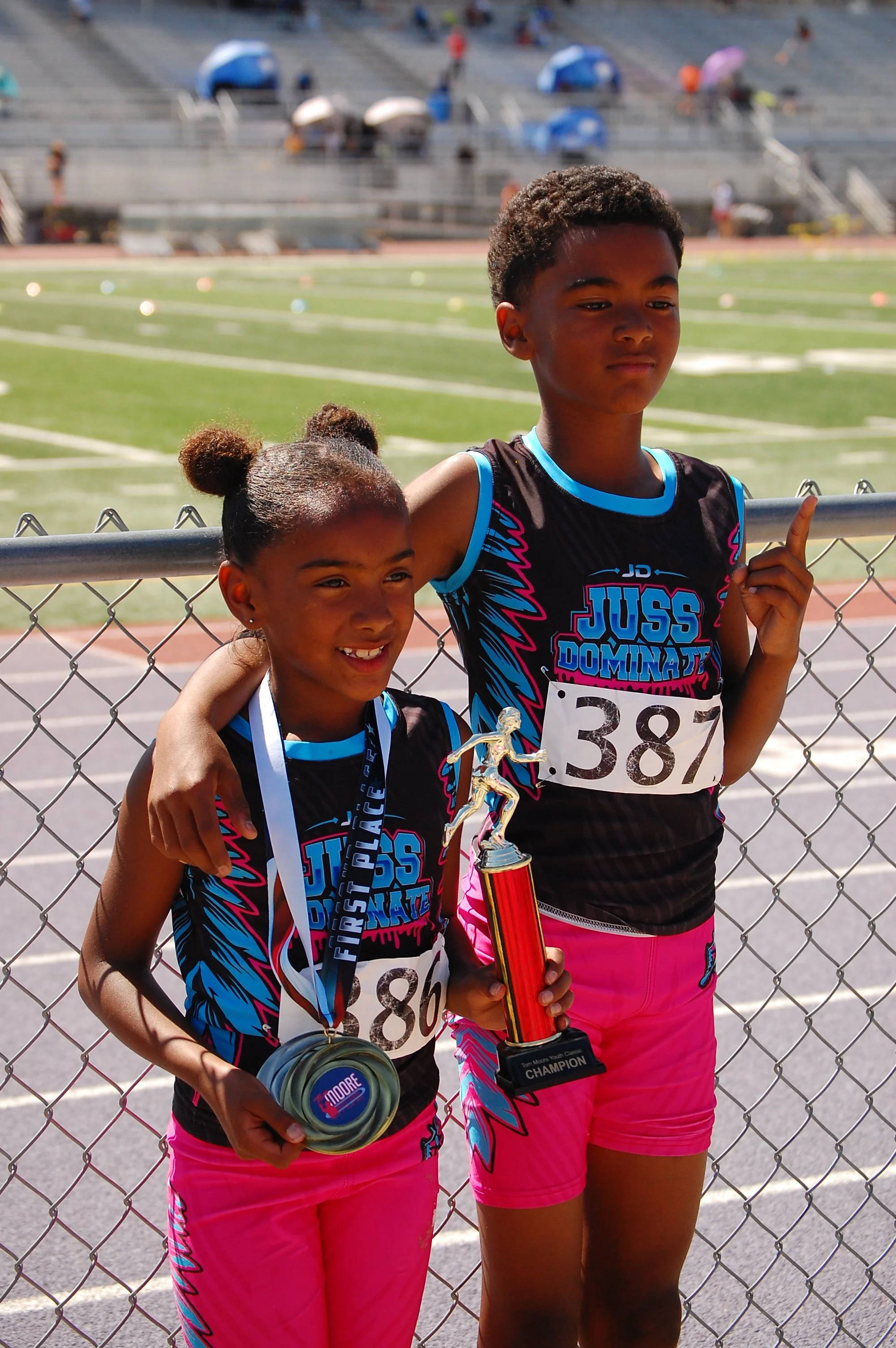 Two young girls in athletic uniforms standing behind a chain-link fence at a track field, holding trophies and medals, smiling and celebrating their victory.