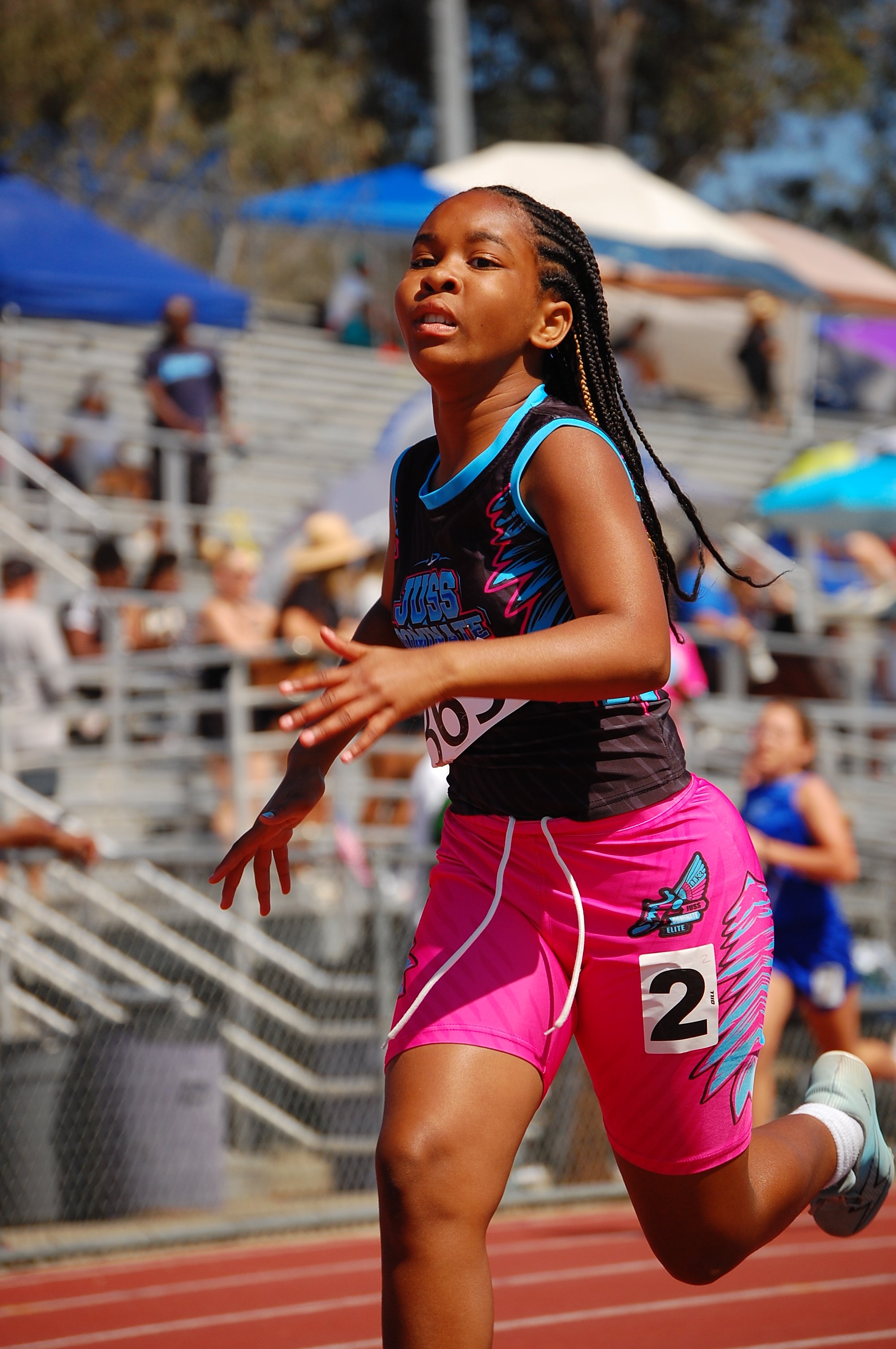 A young female athlete running on a track during a race, wearing pink shorts and a black sleeveless top with blue accents, with a race number 2 on her shorts. She is focused and in motion, surrounded by spectators and other participants in the backgr
