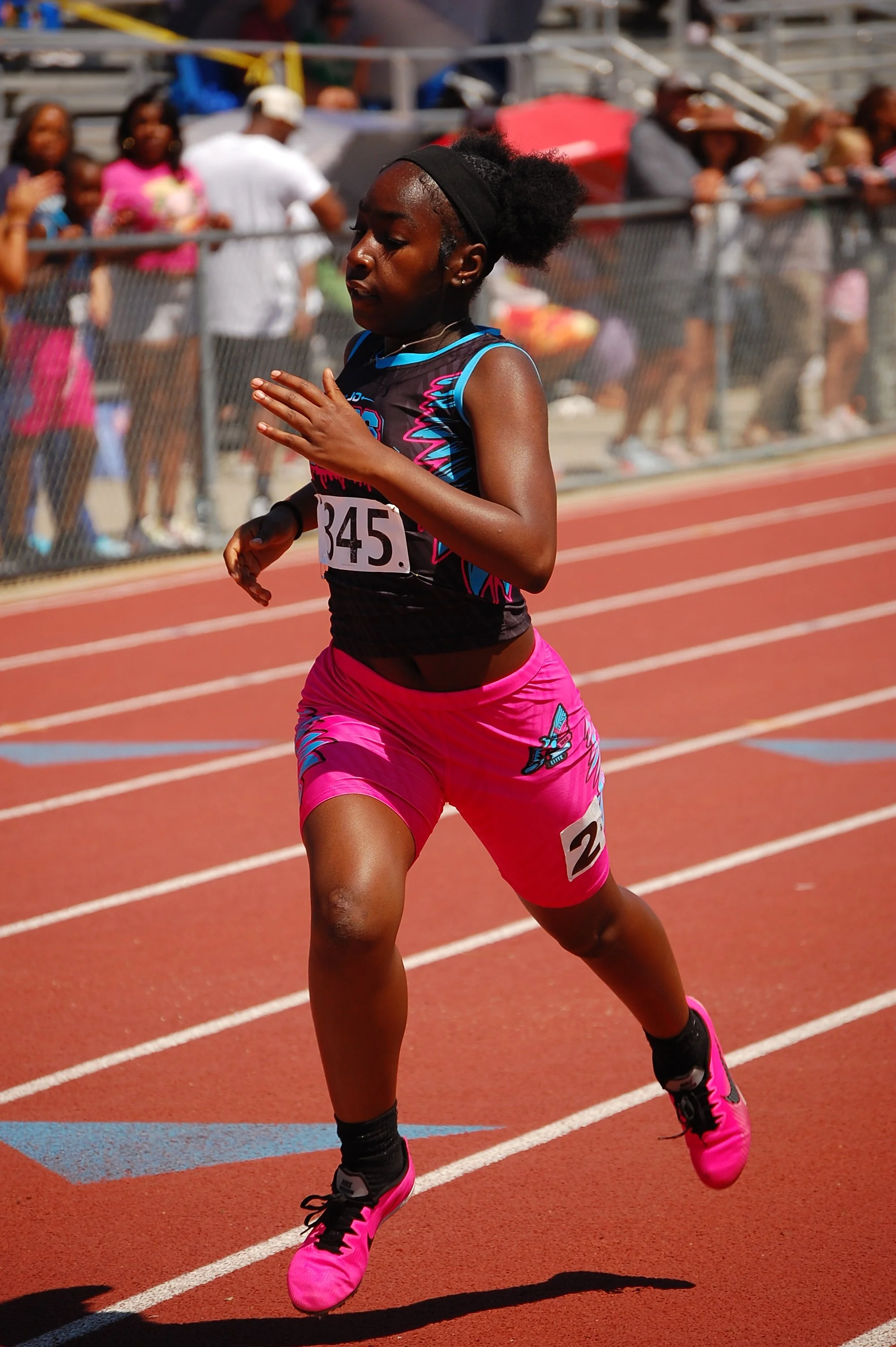 A female athlete running on a track during a race, wearing a black and pink athletic outfit, with a crowd of spectators behind a fence in the background.