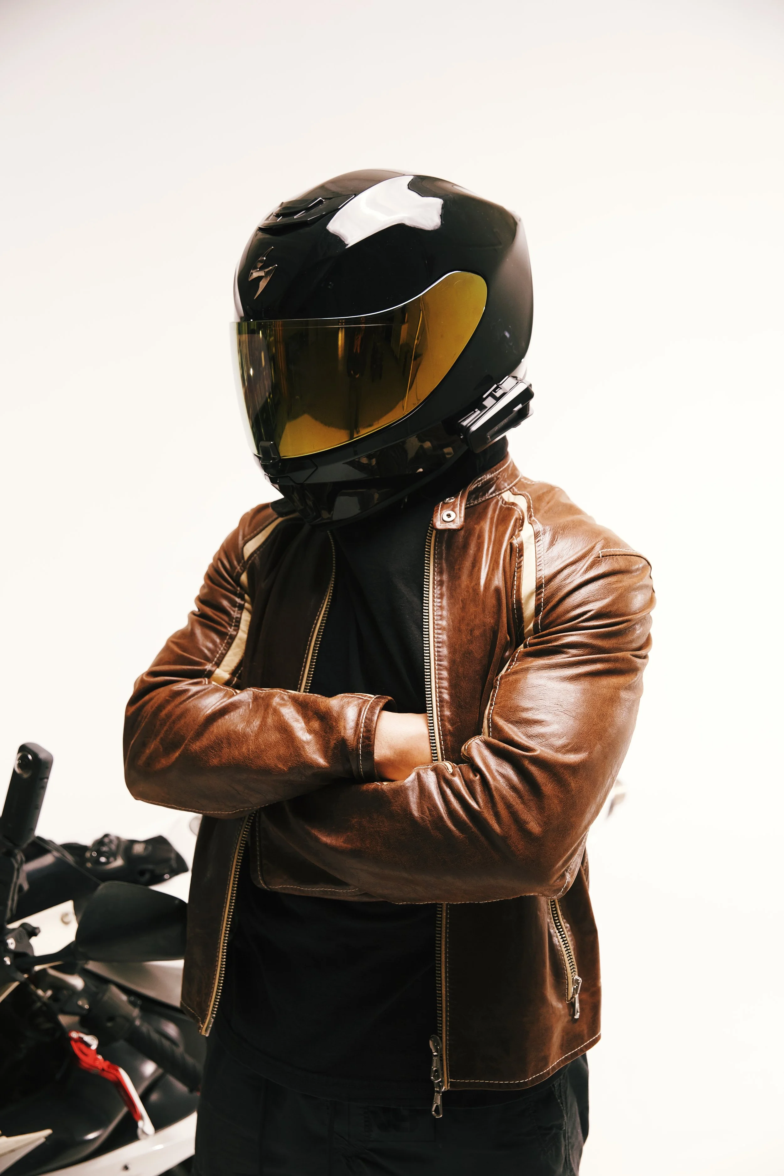 Person wearing a black motorcycle helmet with yellow visor, brown leather jacket, and black shirt, standing with arms crossed next to a motorcycle.