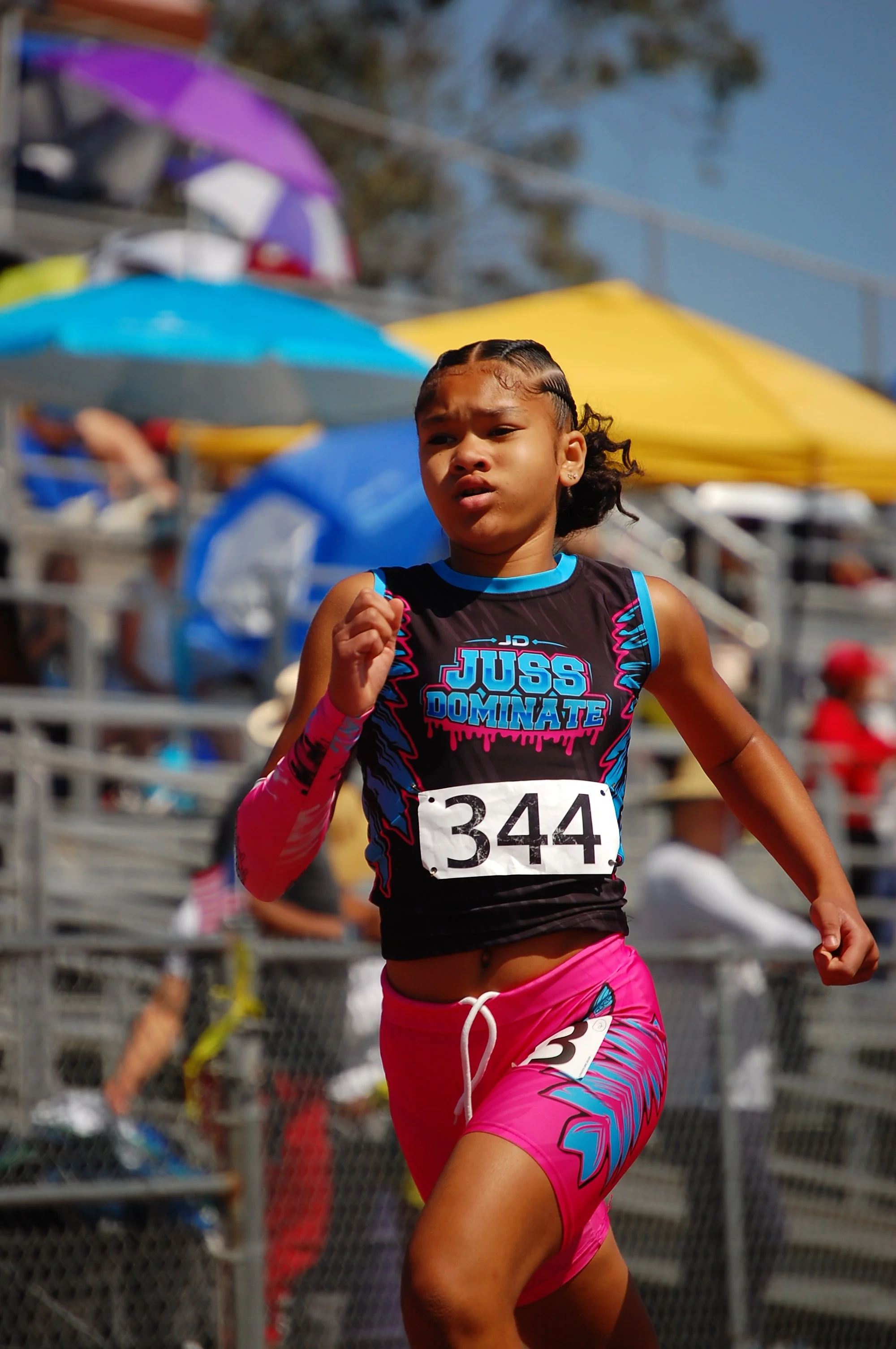 A young girl running in a race during a sporting event, wearing a black and pink athletic uniform with the words "Juss Dominate" on her jersey and the number 344 on her bib, with a determined expression on her face. In the background, there are color
