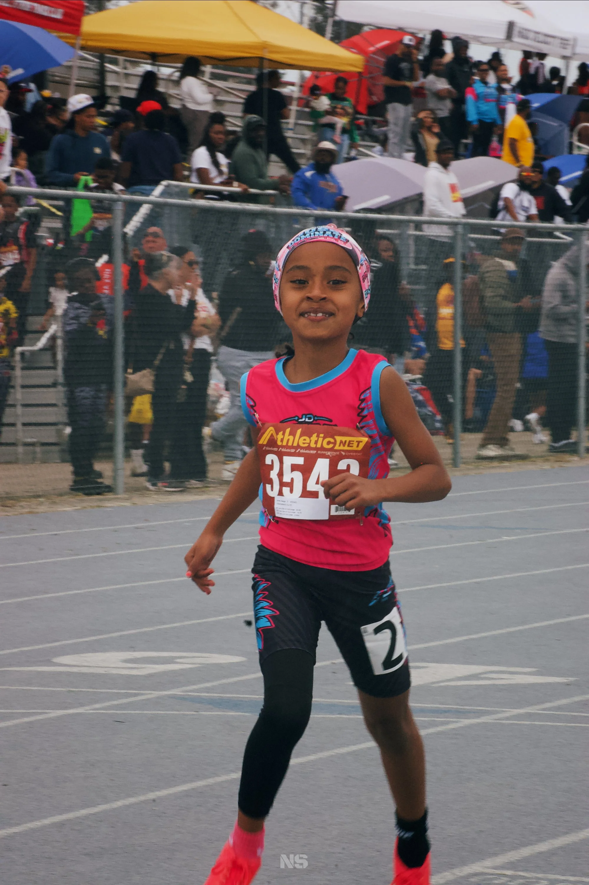 A young girl running in a race at a track event, wearing a pink and blue athletic outfit with a race bib numbered 3543, pink shoes, and a colorful headband. There are spectators and other participants in the background, some holding umbrellas, at an 