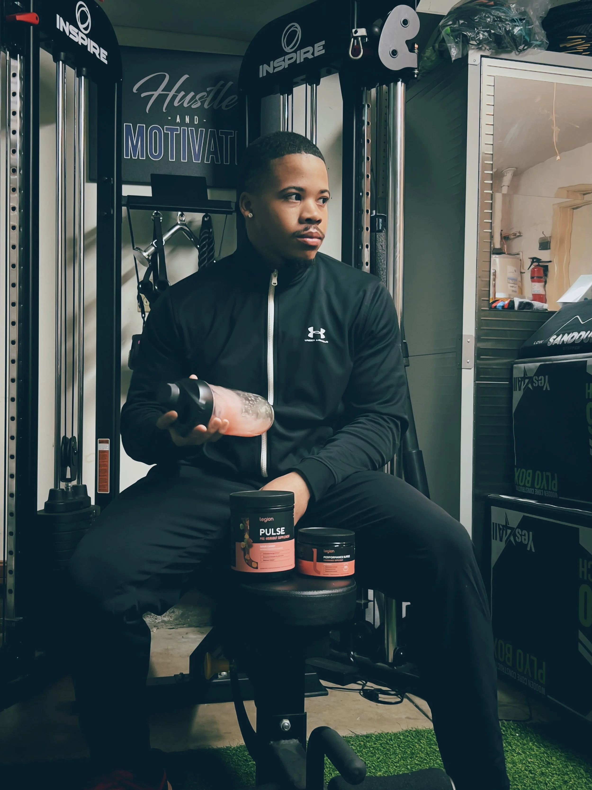 A person sitting on a workout bench holding a water bottle, surrounded by fitness equipment and supplement containers at a gym with motivational signs.