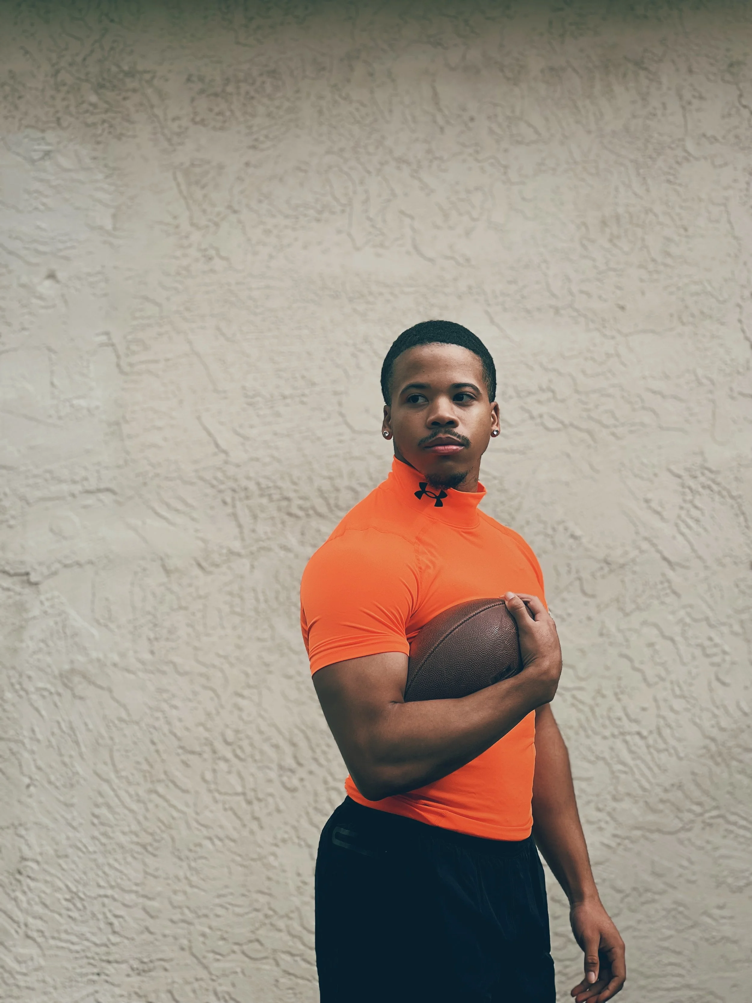 A young man wearing an orange athletic shirt holding a football against a textured beige wall.