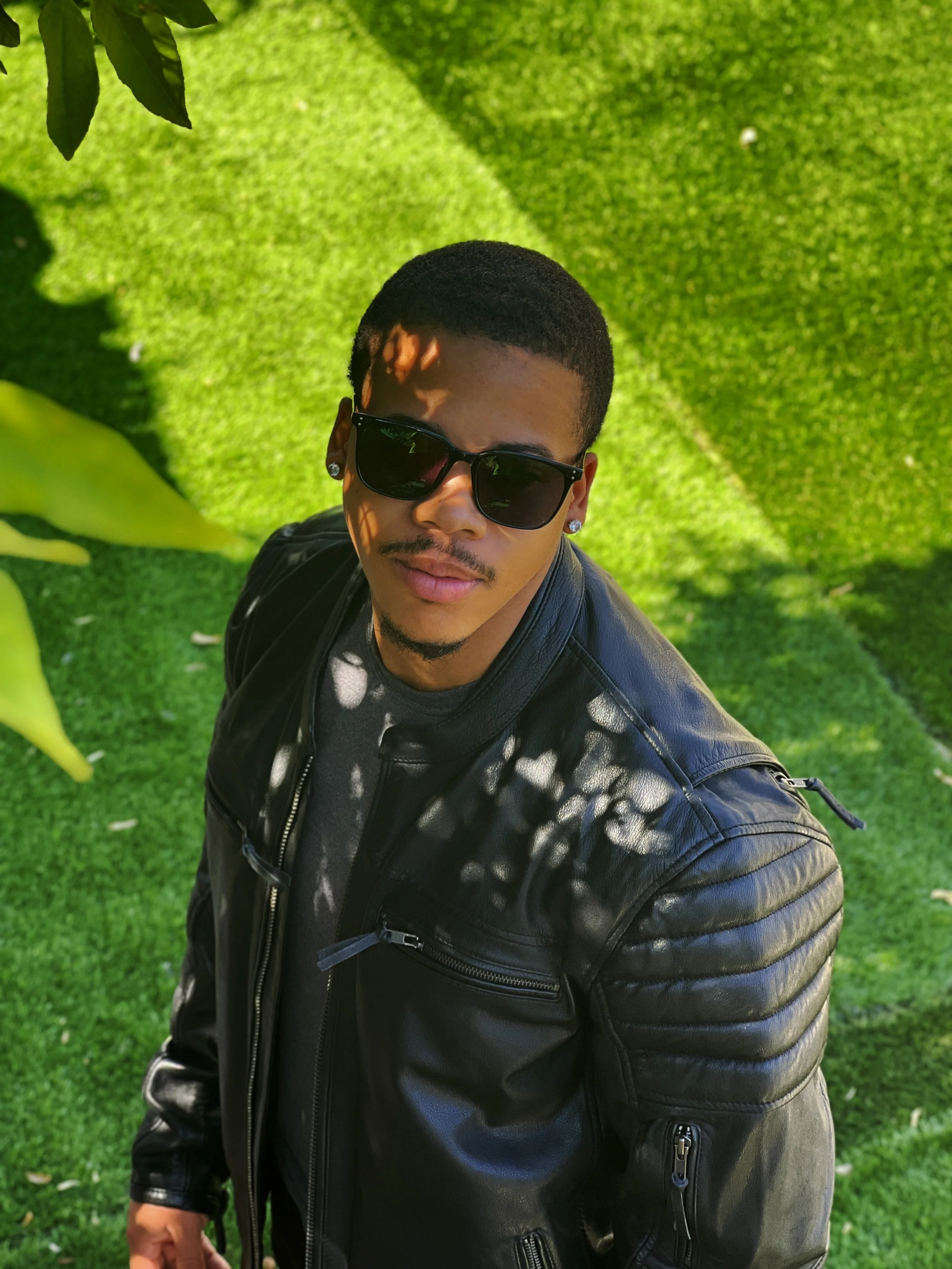 Young man with short hair, wearing sunglasses, earrings, a black leather jacket, and a dark T-shirt, standing outdoors on a bright, sunny day.