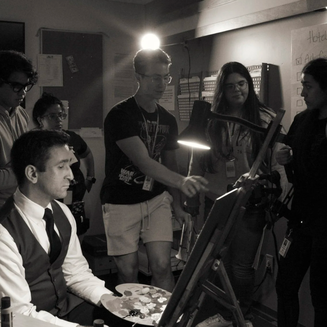 A group of people in a dimly lit room, with one person painting on a canvas and others watching, some using a laptop or holding clipboards.