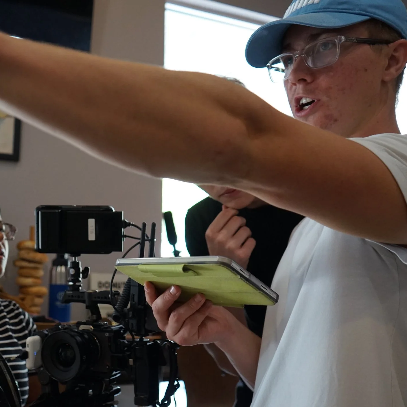 Young man with glasses, wearing a baseball cap, using a tablet while on a film set with camera equipment, other people in the background.