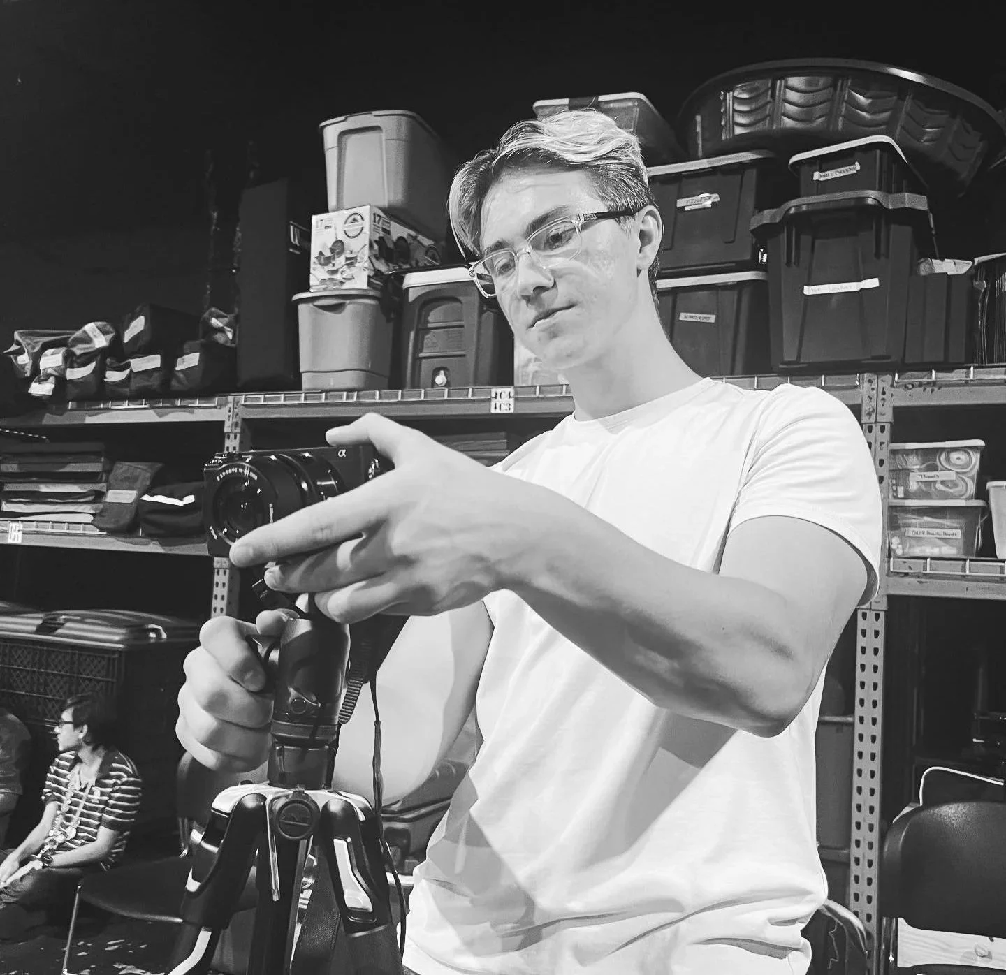 A young man with glasses in a white t-shirt adjusting a camera on a tripod in a storage room or workshop, with shelves filled with storage bins and tools in the background.