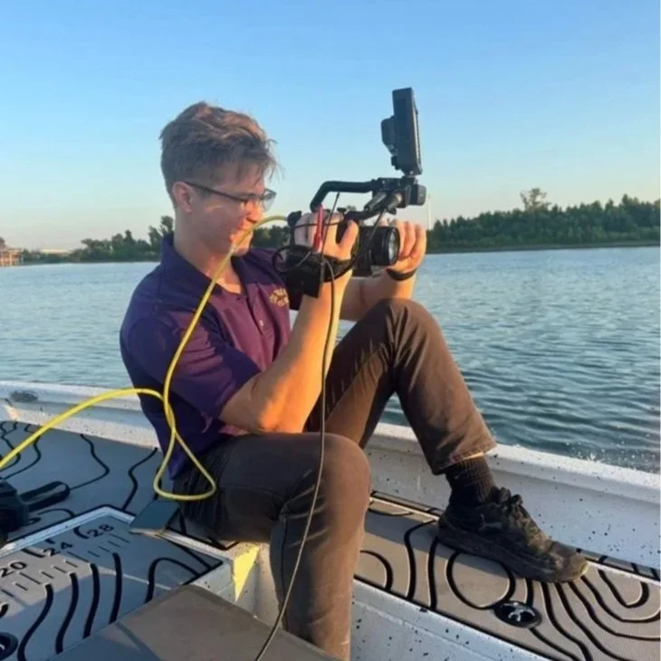 A person on a boat filming with a camera mounted on a stabilizer, with a lake and trees in the background during daytime.