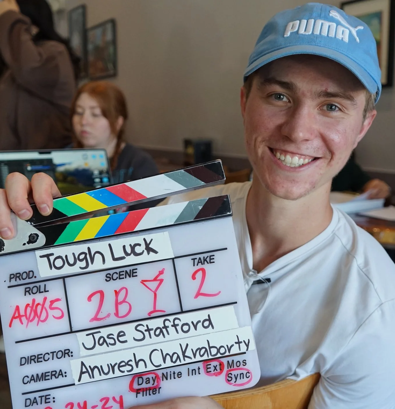 A young man smiling and holding a clapperboard with the text "Tough Luck" written on it, in a film or TV production setting.