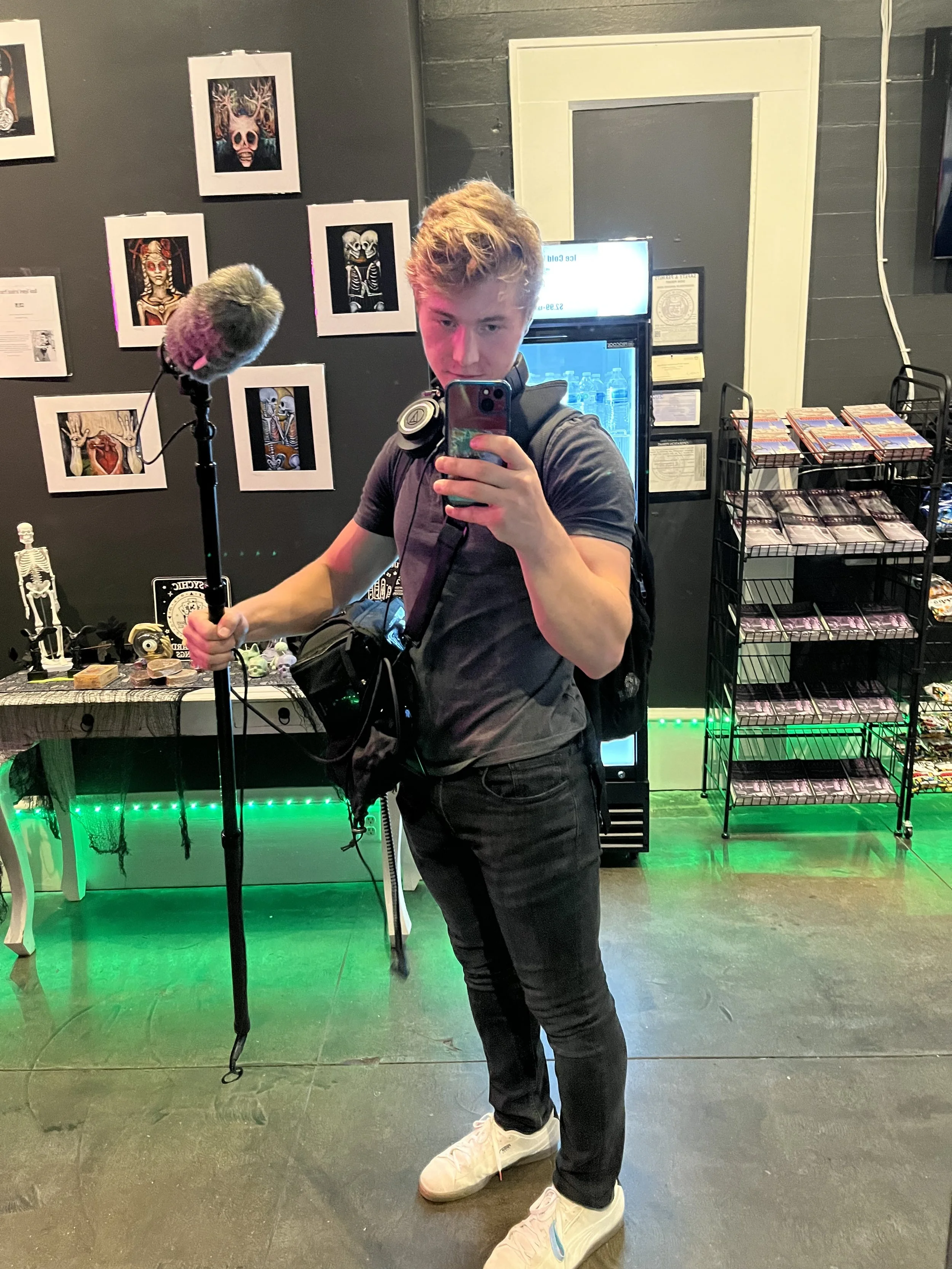 Young man with curly red hair taking a mirror selfie in a tattoo or art shop, holding a microphone windscreen, with a backpack and headphones, surrounded by artwork and display racks.