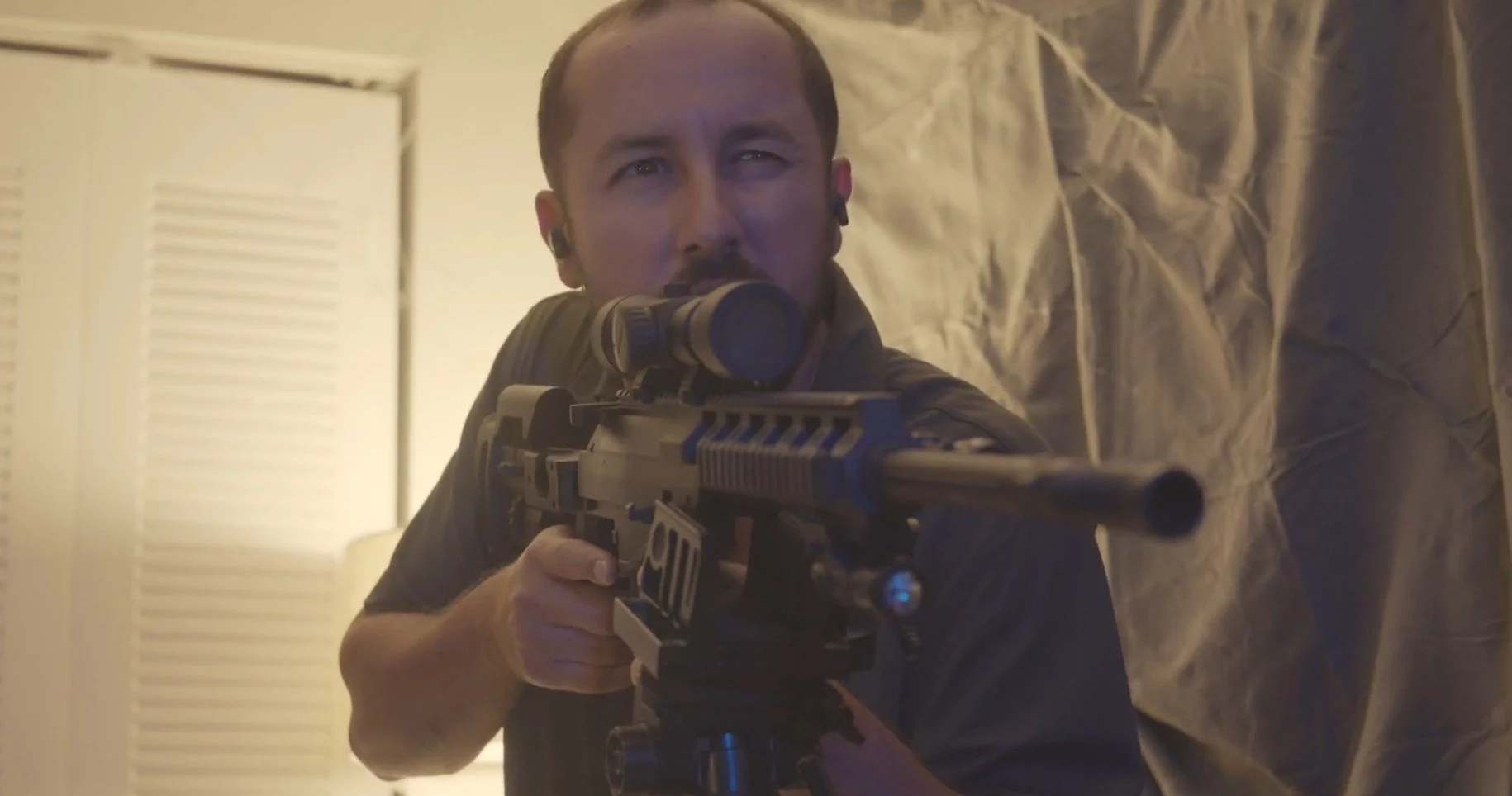 Man aiming a scoped rifle in a room with beige walls and a door.