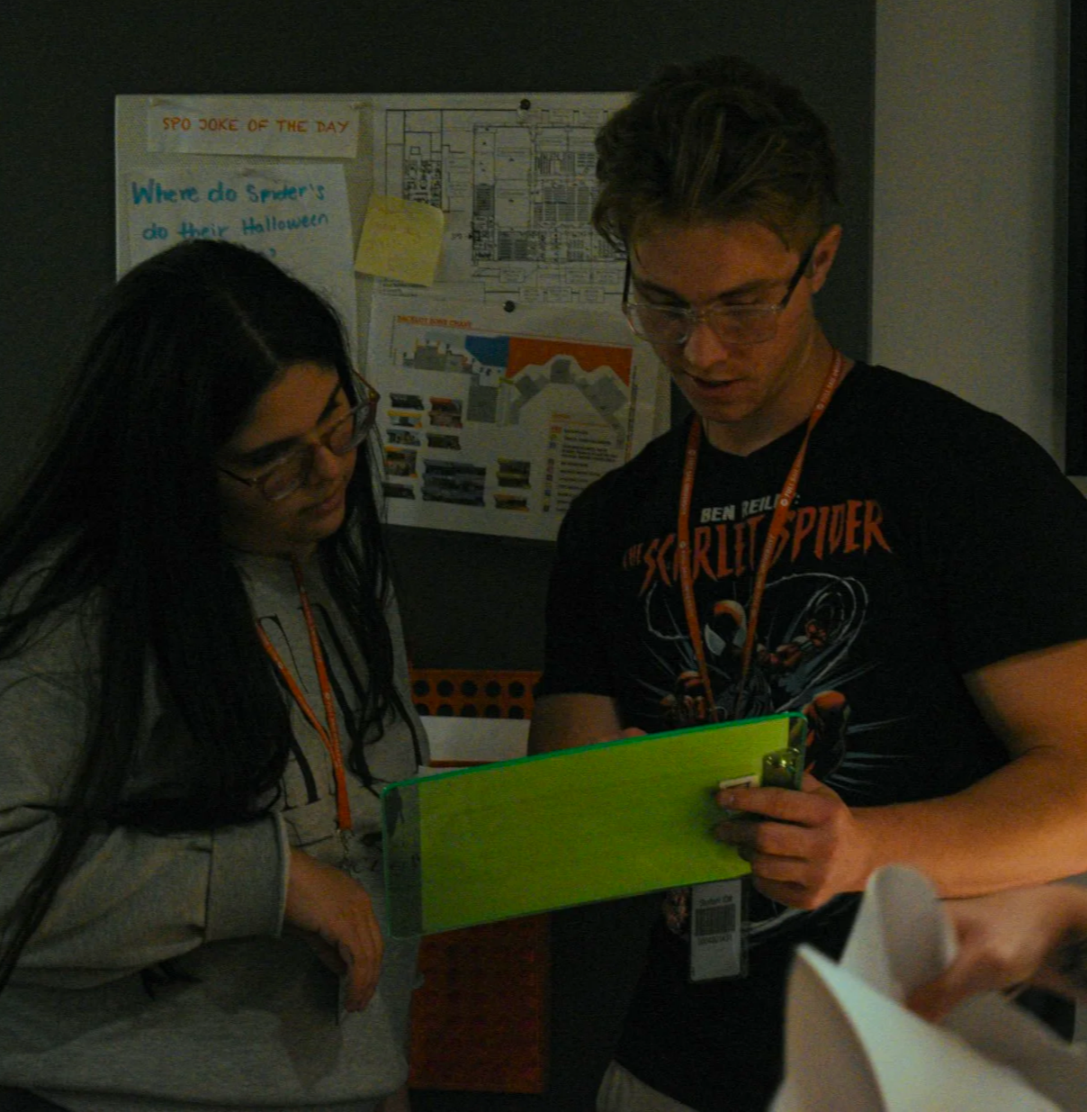 Two people, a woman with long dark hair and a man with light hair, both wearing glasses and black lanyards, looking at a green folder or tablet in a dark indoor setting. There are posters and diagrams on the wall behind them.