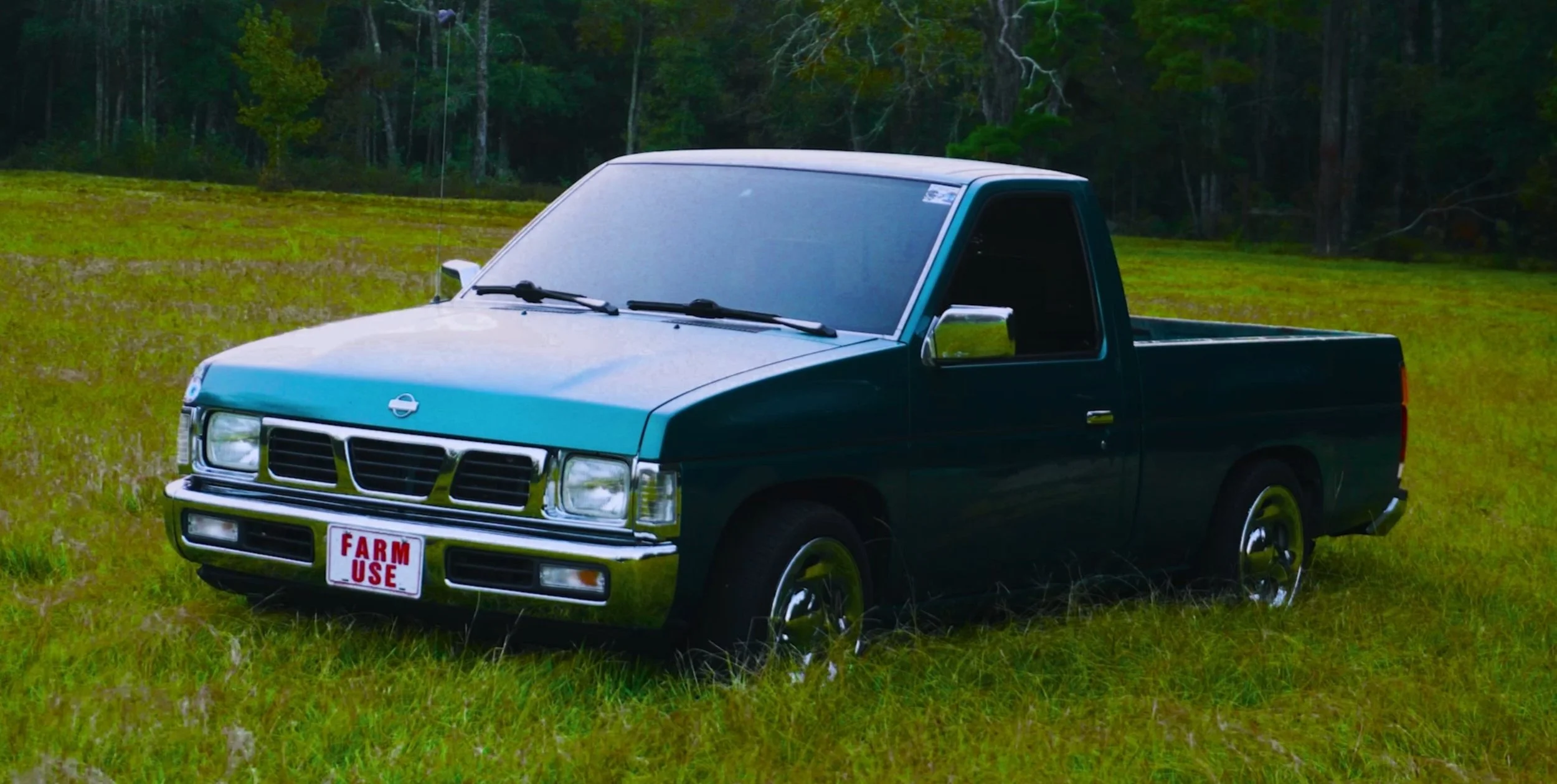 A vintage black and teal pickup truck with a 'Farm Use' license plate parked on grass in a rural area with trees in the background.