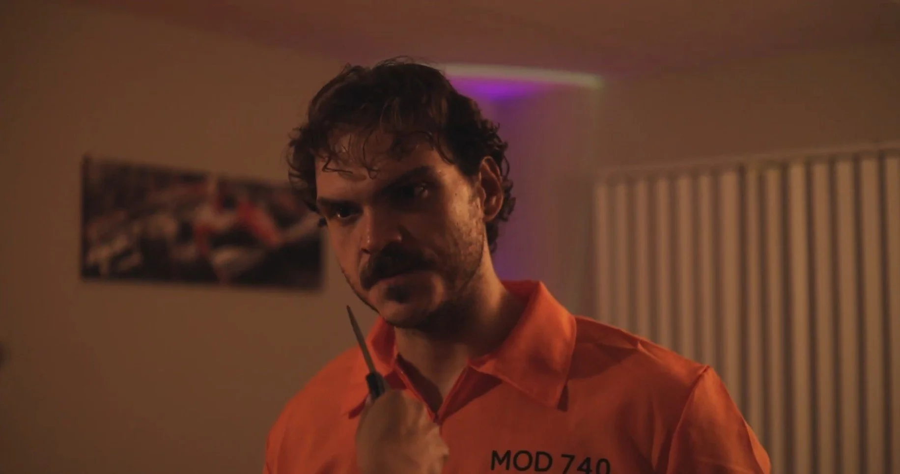 A man with a mustache and curly hair wearing an orange prison jumpsuit with 'MOD 740' on it, holding a small object close to his face in a dimly lit room.