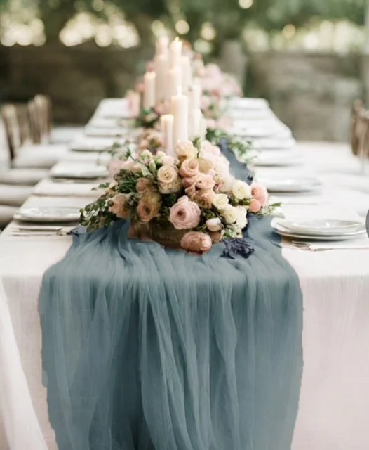 A long dining table decorated with a blue tulle runner and floral centerpiece, set outdoors with candles.