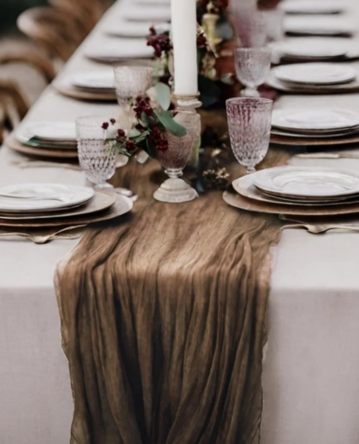Elegant dinner table with white tablecloth, rustic wooden runner, pink textured glasses, white plates, and centerpiece featuring pink and burgundy flowers and greenery with a tall white candle.