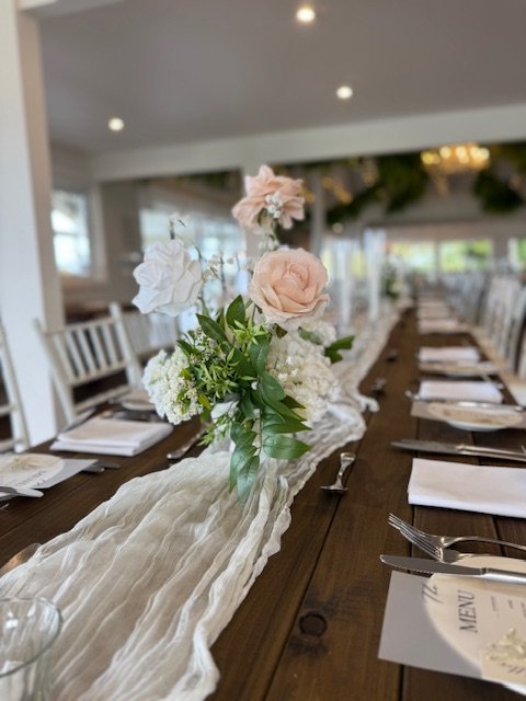 A long wooden banquet table decorated with a white floral centerpiece and a light fabric runner, set with plates, napkins, and utensils in a well-lit dining area.