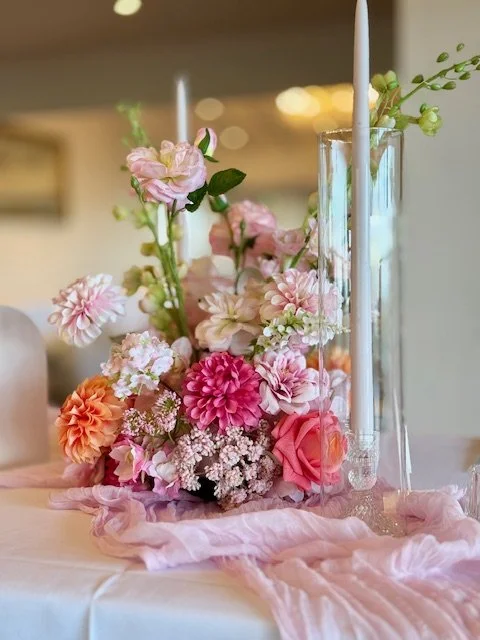 A floral centerpiece with pink, white, and peach flowers and two white candles in glass vases on a table draped with pink fabric.