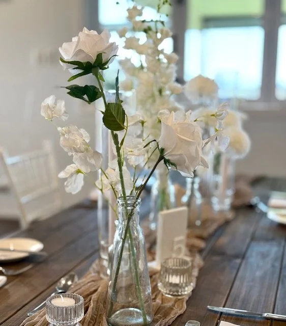 White roses and hydrangeas in glass vases on a wooden dining table decorated with a burlap table runner, with candles and place settings visible in the background.