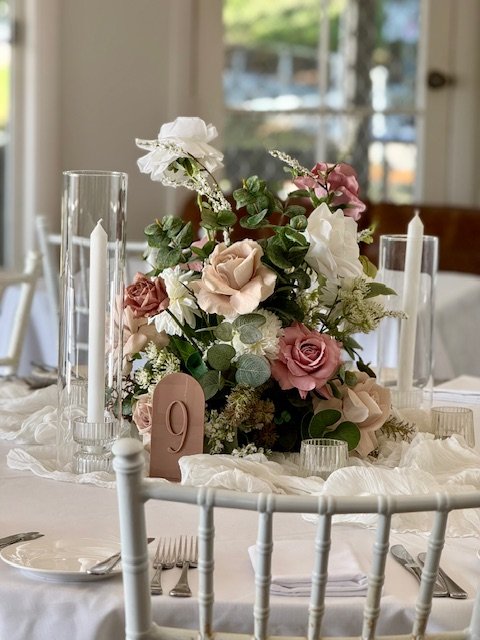 Elegant table centerpiece with pink, white, and blush flowers, surrounded by tall glass candle holders, set on a white tablecloth with silverware and a table number '9' card.