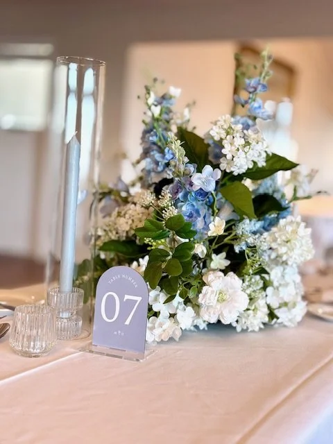 Table centerpiece with white and blue flowers, a blue candle in a glass holder, and a table number sign reading '07' at a decorated event.