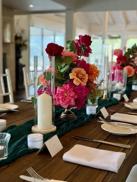 Table centerpiece with pink and orange flowers, green foliage, and a large white candle in a glass holder, arranged on a green table runner at a formal dining table.