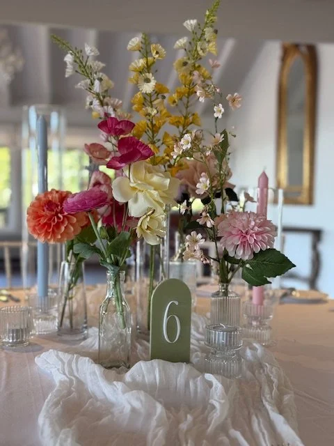 Table centerpiece with mixed flowers in glass jars and a table number 6 card, on a white tablecloth in a decorated room.