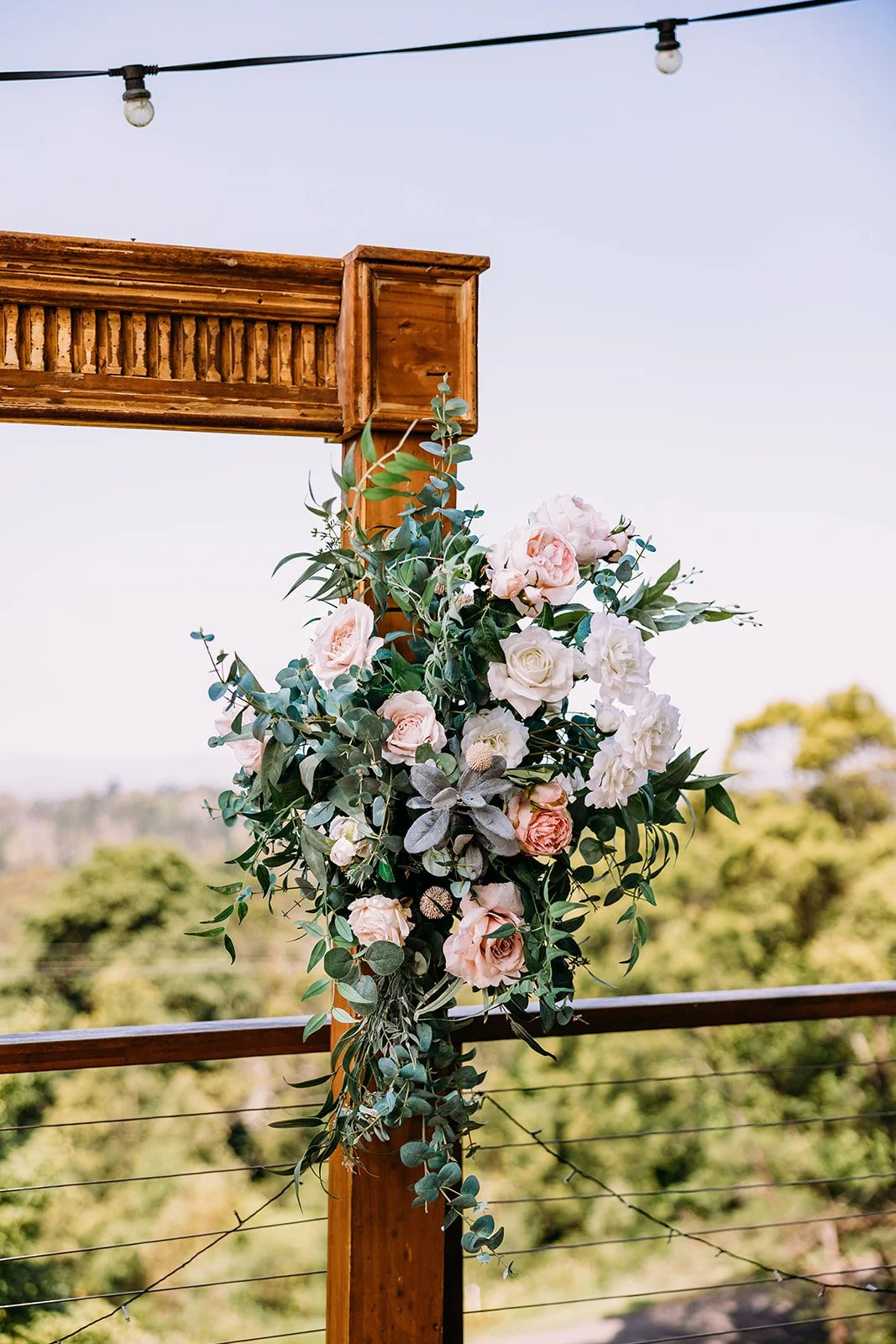 A floral arrangement with pink and white flowers and green foliage hanging on a wooden post at an outdoor event, with string lights overhead and a green landscape in the background.