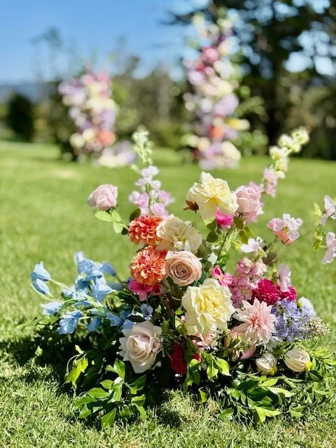 Colorful floral arrangement on grass with blurred background of other flowers and trees.
