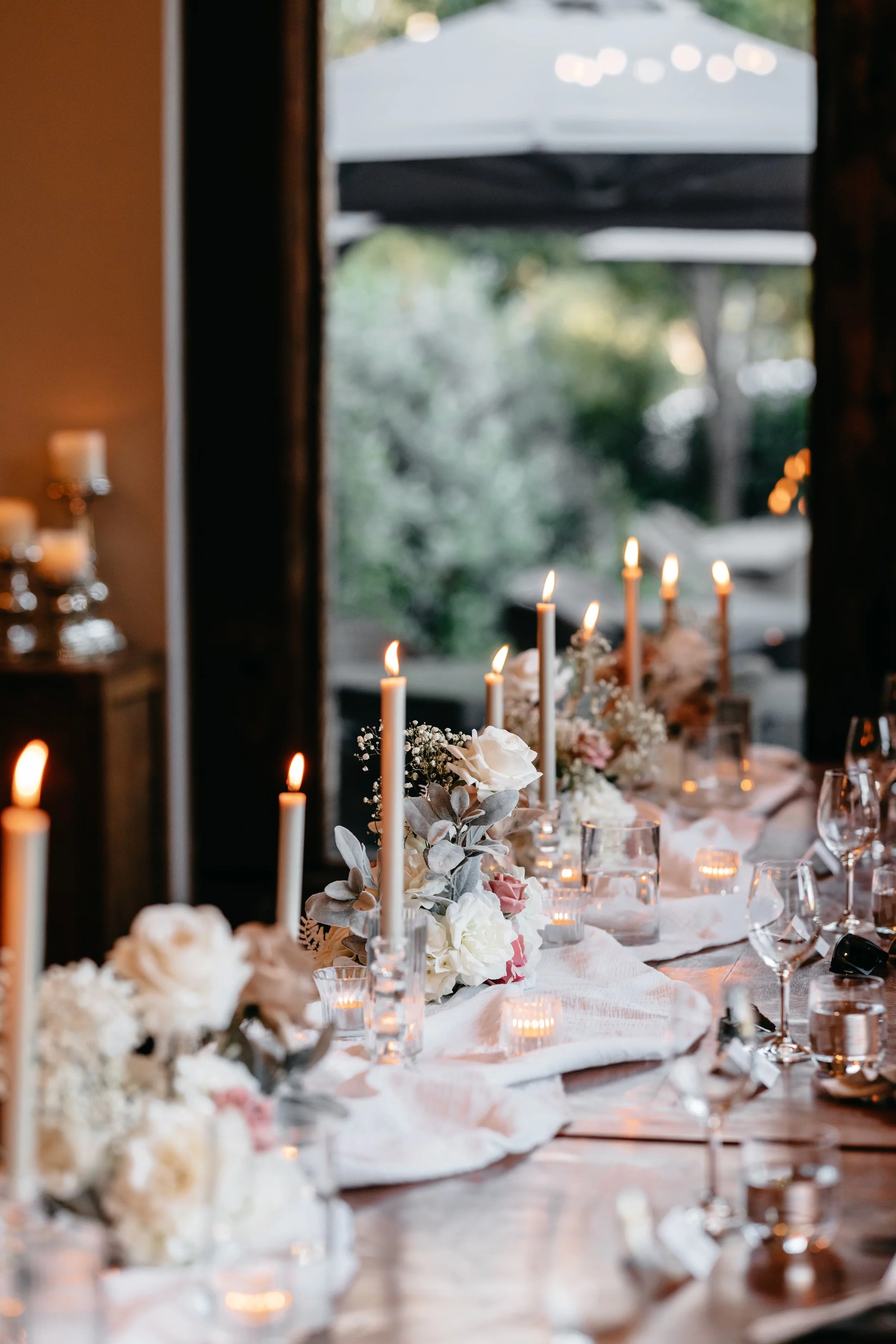 A long dining table decorated with white and pink flowers, candles, and glassware in a softly lit room.