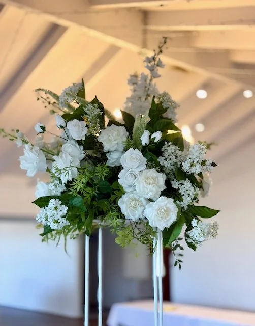 A white floral arrangement with roses and greenery in a silver vase against a blurred indoor background.