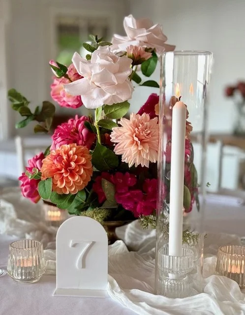 A floral centerpiece with pink, peach, and white flowers in a glass vase, a white candle in a glass holder, and a white number 7 table marker on a table covered with white cloth. Small candles are also visible around the arrangement.