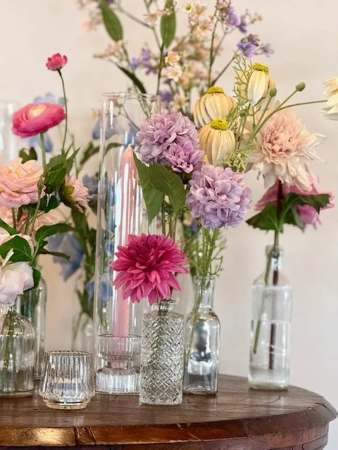 Various fresh flowers in glass vases arranged on a wooden surface, including pink, purple, and cream-colored blooms.