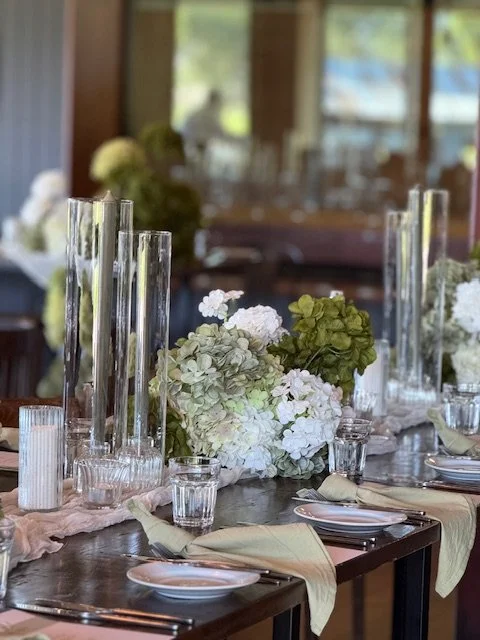 Elegant dining table decorated with white and green flowers, tall glass candle holders, and tableware for a formal event, set inside a bright room with large windows.