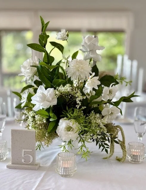 Elegant white floral centerpiece on a dining table with candle holders and a table number sign.