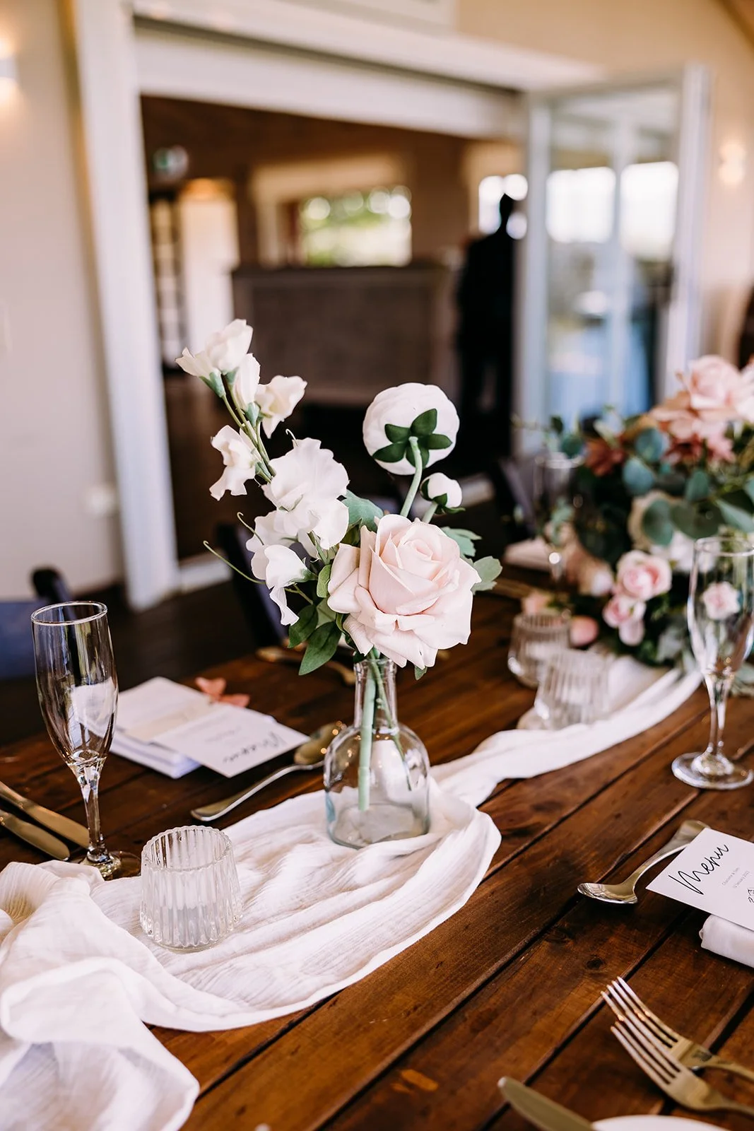 A wedding table centerpiece featuring a clear glass bottle with pink roses, white flowers, and greenery, arranged on a white runner with glassware, silverware, and place cards.