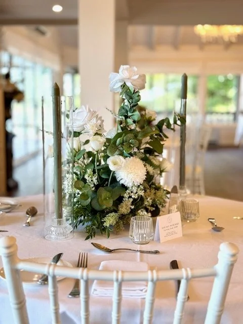 Elegant round table at a wedding reception with a white floral centerpiece, candles, glassware, and place settings.