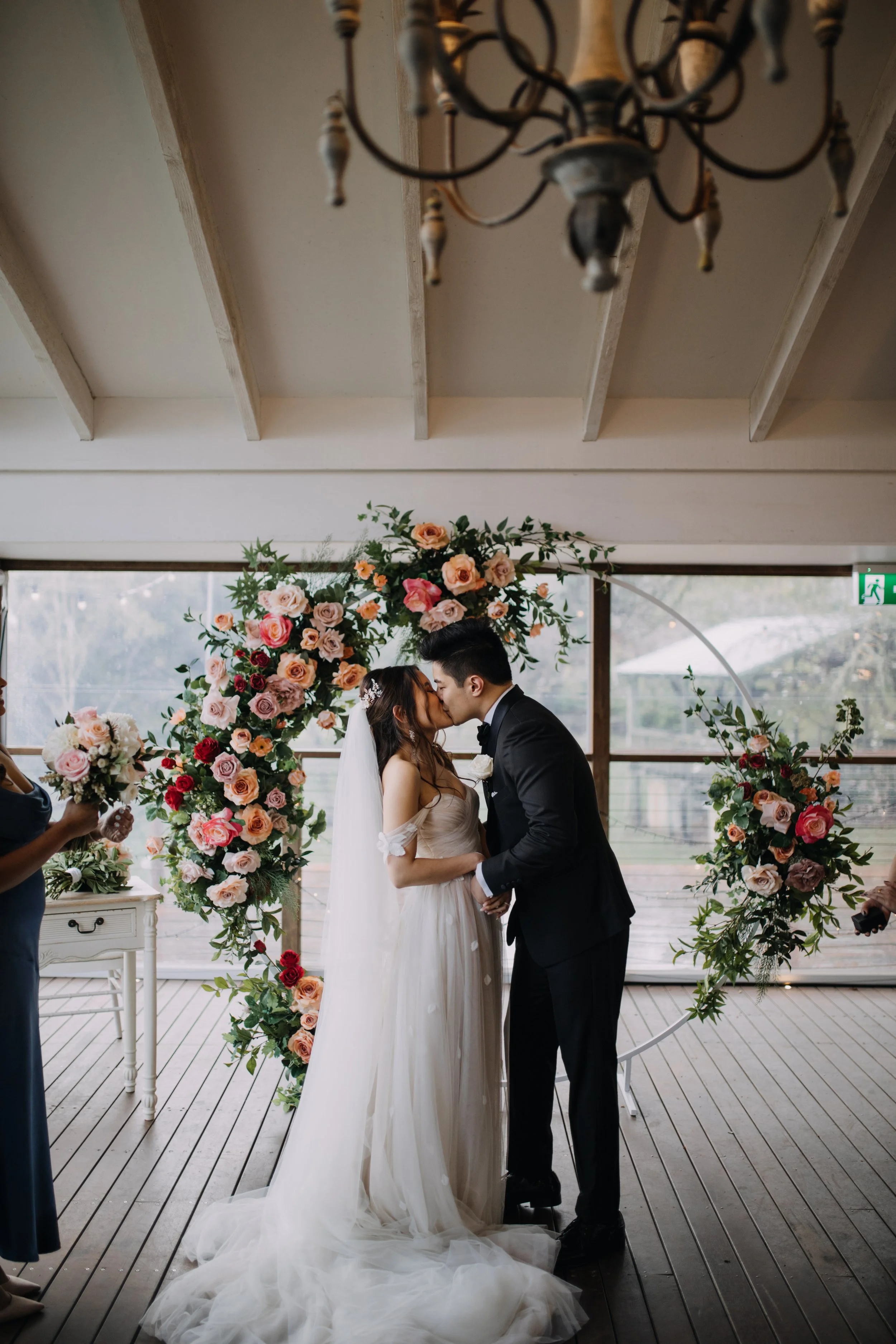 A bride and groom kissing during their wedding ceremony, standing in front of a large floral arch with pink and red roses, inside a venue with wooden floors and large windows.