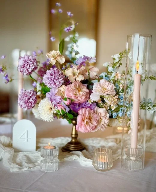Elegant table centerpiece featuring a bouquet of pink, purple, and white flowers in a bronze vase, surrounded by candles in glass holders on a white tablecloth.