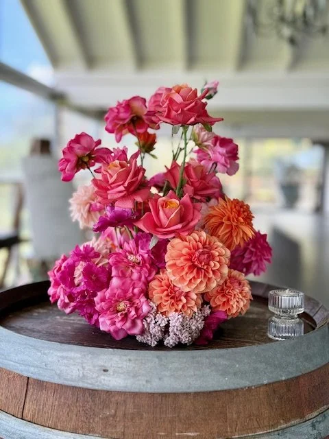 A bouquet of colorful flowers, including pink, peach, and purple blossoms, arranged on a wooden barrel with a small glass container nearby.