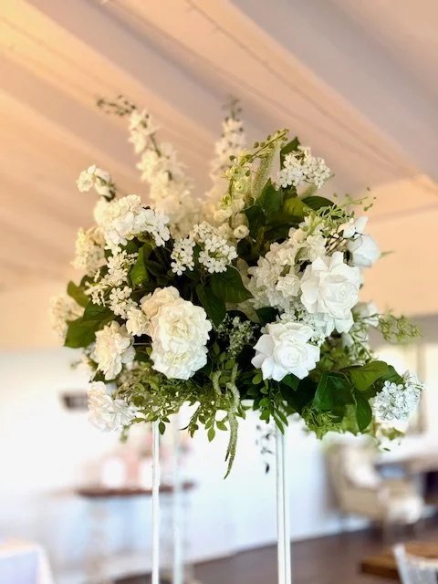 White flower arrangement with roses, hydrangeas, and greenery in a clear glass vase.