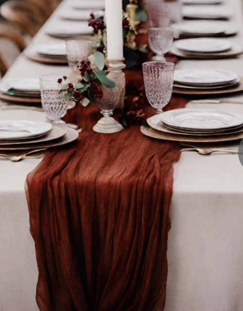 A long dining table set with plates, glasses, and cutlery, decorated with a rust-colored table runner, tall white candle, and floral arrangements with greenery and dark red berries.