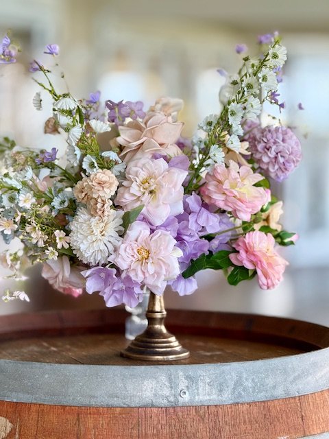 A floral arrangement with pastel-colored flowers including pink, purple, and white, placed in a decorative metallic vase on a wooden surface.