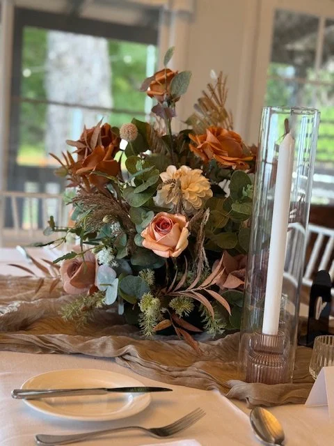 A floral centerpiece with pink roses, white flowers, and greenery on a dining table. There's a tall glass candle holder with a white candle beside it. The background shows a window with trees outside.