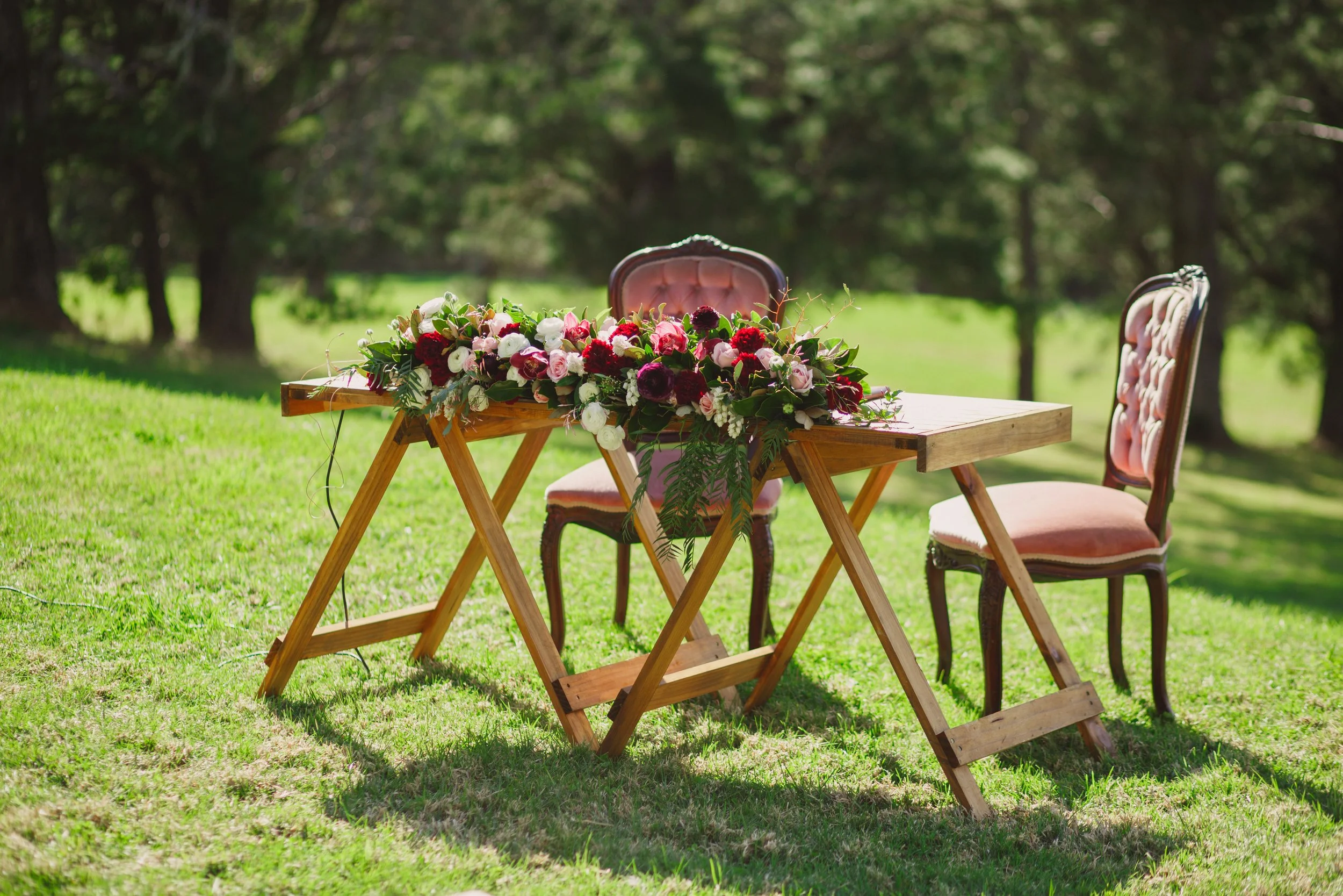 A wooden table decorated with a colorful flower arrangement, set outdoors on grass with trees in the background, accompanied by two vintage pink upholstered chairs.