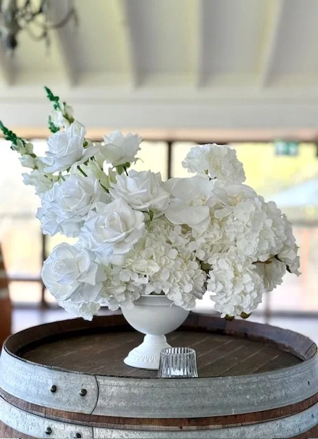 A white flower arrangement in a white ceramic vase on top of a wooden barrel, with a small glass object in front, inside a room with a window in the background.