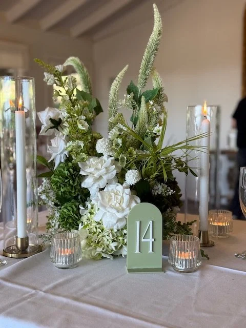 Elegant table centerpiece with white flowers, greenery, tall white candles in glass holders, and a table number 14 at a formal event.