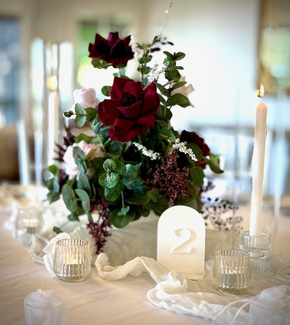 A floral centerpiece with red and white roses, greenery, and dark purple accents on a table with glass candle holders, a white card with the number 2, and a lit white candle.